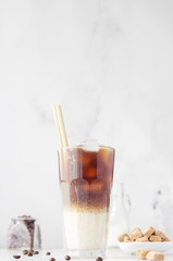 Cold coffee with milk and ice cubes in a tall glass with bamboo straw, light grey background. Selective focus. 