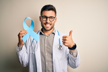 Naklejka premium Young professional doctor man holding colon awareness blue ribbon over isolated background happy with big smile doing ok sign, thumb up with fingers, excellent sign