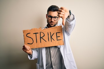 Young handsome doctor man protesting holding cardboard with strike message with angry face, negative sign showing dislike with thumbs down, rejection concept