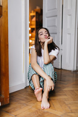 Barefooted cute girl sitting on the floor at home and fooling around. Indoor portrait of brunette young lady posing in her apartment with kissing face expression.