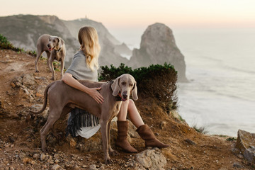 Blond Woman sitting on a sea cliff in Portugal, looking out to the horizon, with her Weimaraner dog .