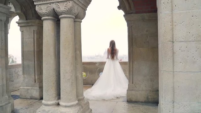 young sophisticated, luxurious bride girl stands between the arches on the terrace or balcony of an ancient castle. Old architectural building of the city. background of the landscape  Budapest