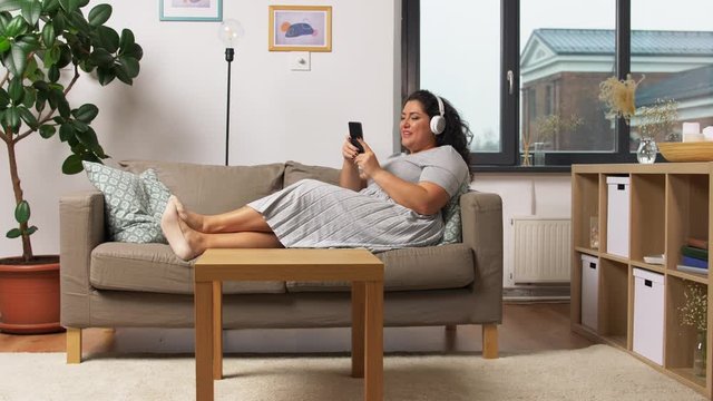 technology, people and leisure concept - happy young woman in headphones listening to music on smartphone on sofa at home