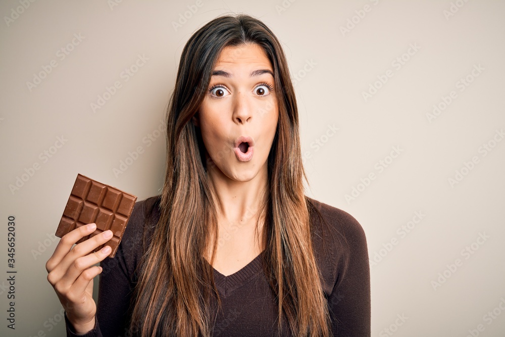 Sticker Young beautiful girl holding sweet bar of chocolate over isolated white background scared in shock with a surprise face, afraid and excited with fear expression