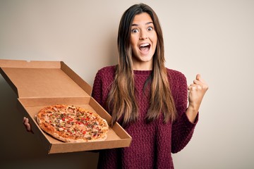Young beautiful girl holding delivery box with Italian pizza standing over white background...