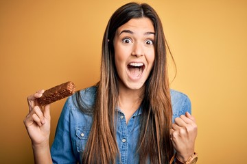 Young beautiful girl holding healthy protein bar standing over isolated yellow background screaming...