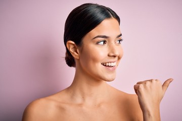 Young beautiful girl standing over isolated pink background pointing and showing with thumb up to the side with happy face smiling