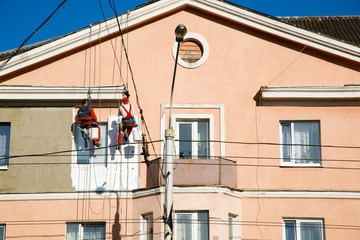 Fototapeta na wymiar Construction workers hanging on rope and renovating building facade at height. Dangerous and risky job of industrial climbers. Plasterers repairing house facade. Reconstruction of building