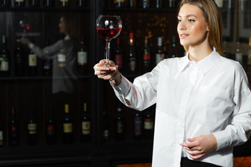 Beautiful blonde young girl sommelier holding up and looking through a glass of wine. Color and bouquet of wine.