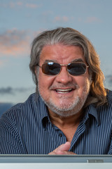 Portrait of a happy latin man when he is standing outside of a window. He is looking down at the camera while leaning the window pane. The sky serves as background. He is wearing sunglasses.