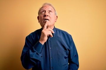 Grey haired senior man wearing casual blue shirt standing over yellow background Thinking concentrated about doubt with finger on chin and looking up wondering