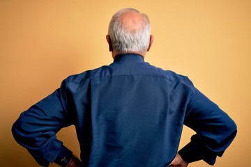 Grey haired senior man wearing casual blue shirt standing over yellow background standing backwards looking away with arms on body - Powered by Adobe
