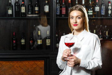 Young beautiful blonde smilling girl sommelier is holding a glass of red wine. Sommelier smelling wine bouquet.