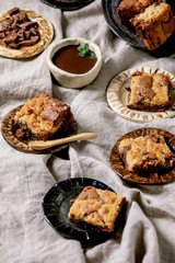 Trend baking Brookies chocolate brownies and cookies homemade cake sliced by squares in different ceramic plates, chocolate sauce, wooden spoons on grey linen table cloth