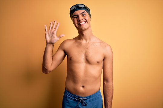 Young handsome man shirtless wearing swimsuit and swim cap over isolated yellow background showing and pointing up with fingers number five while smiling confident and happy.