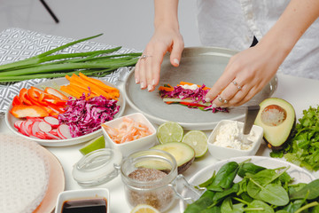 Woman's hands adding red cabbage and cheese to the spring roll on rice paper. Table with spring rolls ingredients, organic and fresh vegetables.