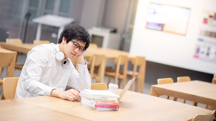 Fototapeta premium Young Asian man university student with glasses and headphones feeling bored while using laptop computer in college library. Doing research and online study class on internet. E-learning concept