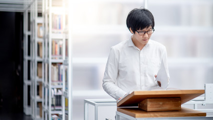 Smart Asian man university student reading recommended book on wooden podium in college library....