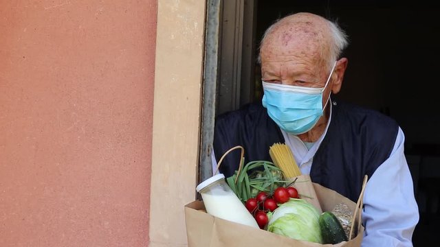 Woman delivering food in paper bag during Covid 19 outbreak. Feme volunteer holding groceries in the house porch. Female volunteer helping disabled old man use smartphone. Food box donation. Social he