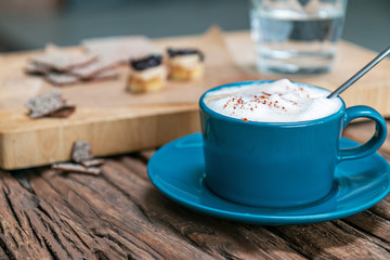 Cappuccino coffee in a turquoise mug on a wooden table against the background of raw desserts and crackers in a low key.