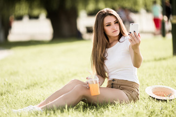Beautiful young woman sitting on grass and using phone in summer park.
