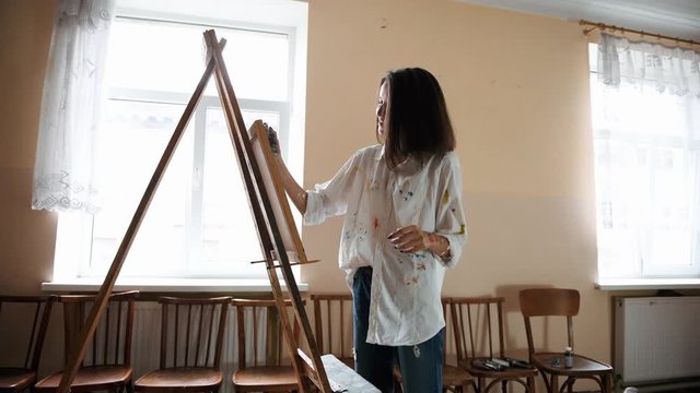 Female painter works with picture by using easel in the studio at daytime. Having good time.