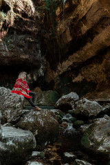Attractive blonde curly young woman wearing red authentic poncho with ethnic pattern, in the beautiful stony gorge or cave 
