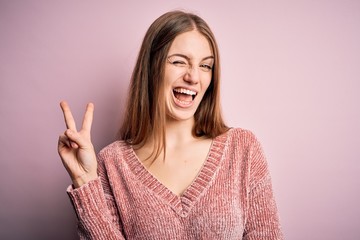 Young beautiful redhead woman wearing casual sweater over isolated pink background smiling with happy face winking at the camera doing victory sign. Number two.