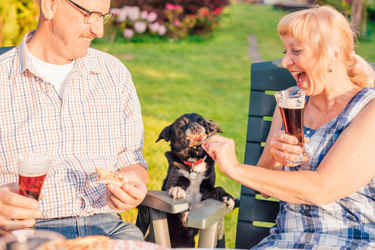 Happy Senior Couple Feeding Dog With Pizza During Backyard Dinner