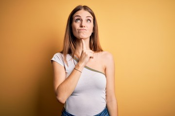 Obraz premium Young beautiful redhead woman wearing casual t-shirt over isolated yellow background Thinking concentrated about doubt with finger on chin and looking up wondering