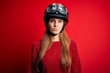 Young beautiful redhead motocyclist woman wearing moto helmet over red background with serious expression on face. Simple and natural looking at the camera.