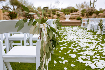 wedding ceremony aisle with white chairs and white flower petals on walkway and eucalyptus leaves tied to chairs