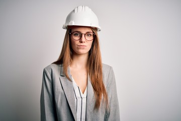 Young beautiful redhead architect woman wearing security helmet over white background with serious expression on face. Simple and natural looking at the camera.