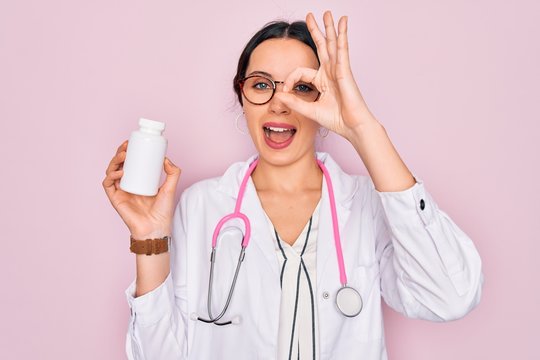 Young beautiful doctor woman with blue eyes wearing stethoscope holding medicine pills with happy face smiling doing ok sign with hand on eye looking through fingers
