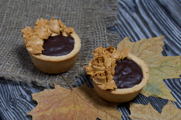 Tartlets with chocolate ganache. On a rough linen cloth among dry maple leaves. Decorated with oil cream flowers. The cream has a caramel color. On brushed pine boards painted black.