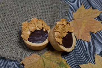 Tartlets with chocolate ganache. On a rough linen cloth among dry maple leaves. Decorated with oil cream flowers. The cream has a caramel color. On brushed pine boards painted black.