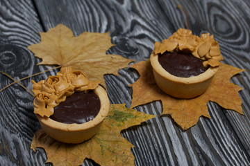 Tartlets with chocolate ganache. On autumn maple leaves. Decorated with oil cream flowers. The cream has a caramel color. On brushed pine boards painted black.
