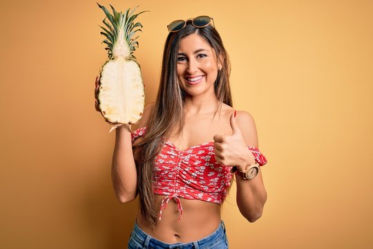 Young beautiful brunette woman holding middle pineapple fruit over isolated yellow background happy with big smile doing ok sign, thumb up with fingers, excellent sign