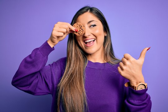 Young beautiful brunette woman holding healthy chocolate cookie over eye pointing and showing with thumb up to the side with happy face smiling