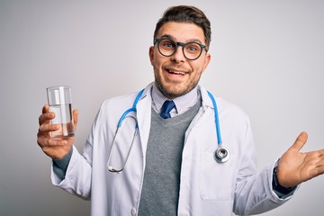 Young doctor man wearing medical coat holding a glass of fresh water over isolated background very happy and excited, winner expression celebrating victory screaming with big smile and raised hands