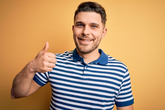 Young man with blue eyes wearing casual striped t-shirt over yellow background doing happy thumbs up gesture with hand. Approving expression looking at the camera showing success.
