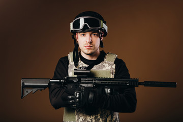 Soldier with gun is standing and looking on camera on brown background. Concept of war. Veterans,...