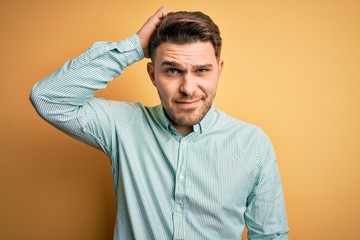 Young business man with blue eyes wearing elegant green shirt over yellow background confuse and wonder about question. Uncertain with doubt, thinking with hand on head. Pensive concept.