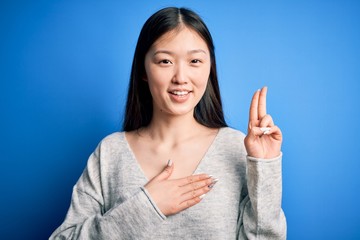 Young beautiful asian woman wearing casual sweater standing over blue isolated background smiling swearing with hand on chest and fingers up, making a loyalty promise oath