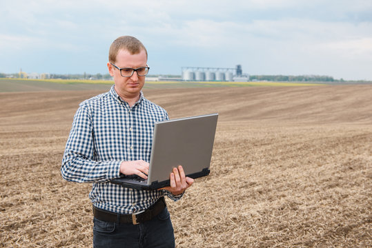 Happy Farmer With Laptop Standing In Wheat Field In Front Of Grain Silo