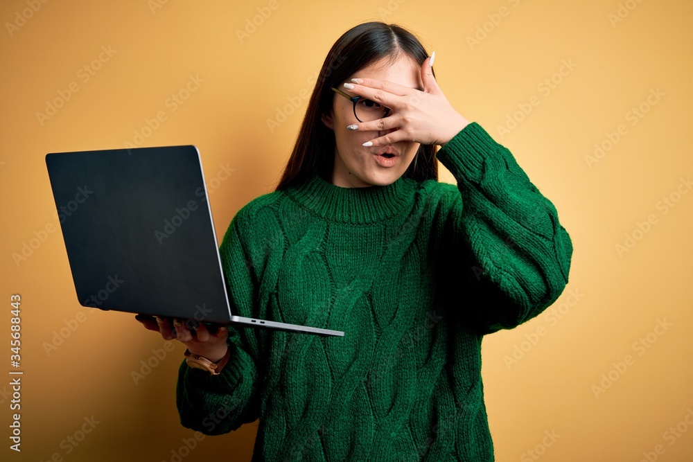 Poster Young asian business woman wearing glasses and working using computer laptop peeking in shock covering face and eyes with hand, looking through fingers with embarrassed expression.