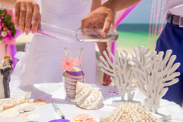 Bride and groom pouring colorful different colored sands into the crystal vase close up during symbolic nautical decor destination wedding marriage ceremony on sandy beach in front of the ocean in Pun