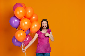 happy young caucasian woman in pink t-shirt pointing at bright colorful air balloons isolated on orange background. birthday party. space for text