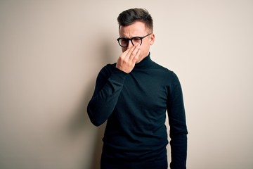Obraz premium Young handsome caucasian man wearing glasses and casual sweater over isolated background smelling something stinky and disgusting, intolerable smell, holding breath with fingers on nose. Bad smell