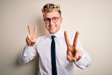 Young handsome caucasian business man wearing golden crown over isolated background smiling looking to the camera showing fingers doing victory sign. Number two.